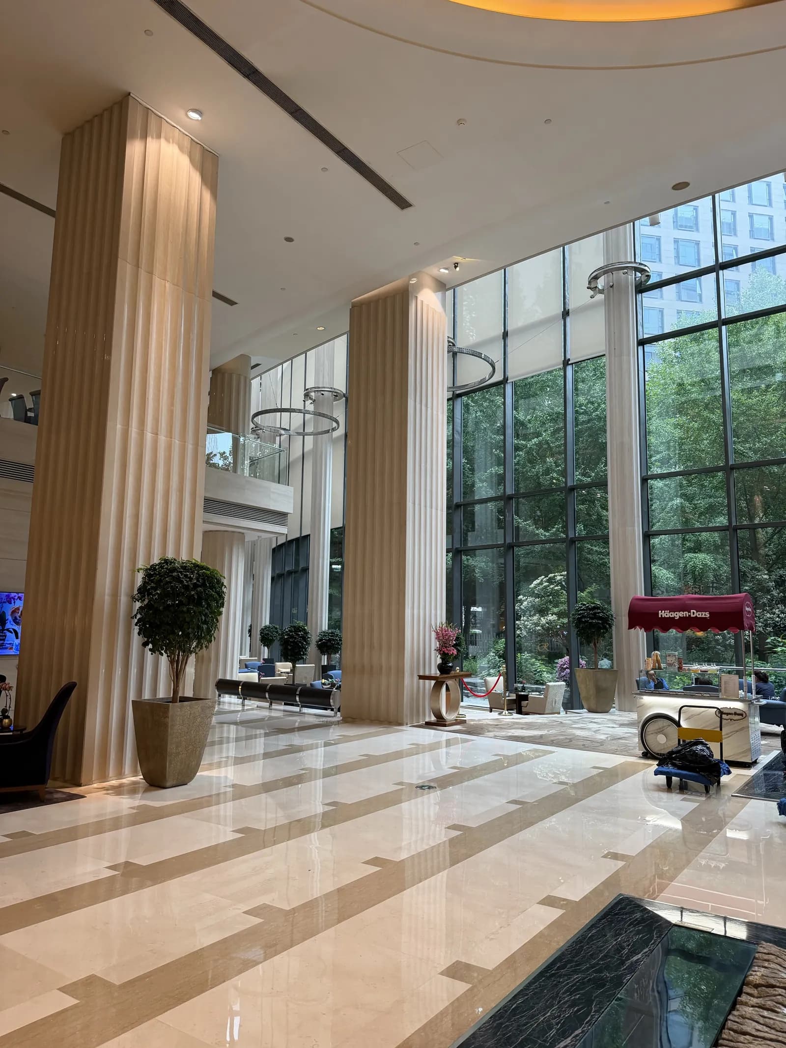 Hohe Hotellobby mit Marmorboden, hellen Säulen und einer Glasfront, hinter der Bäume zu sehen sind