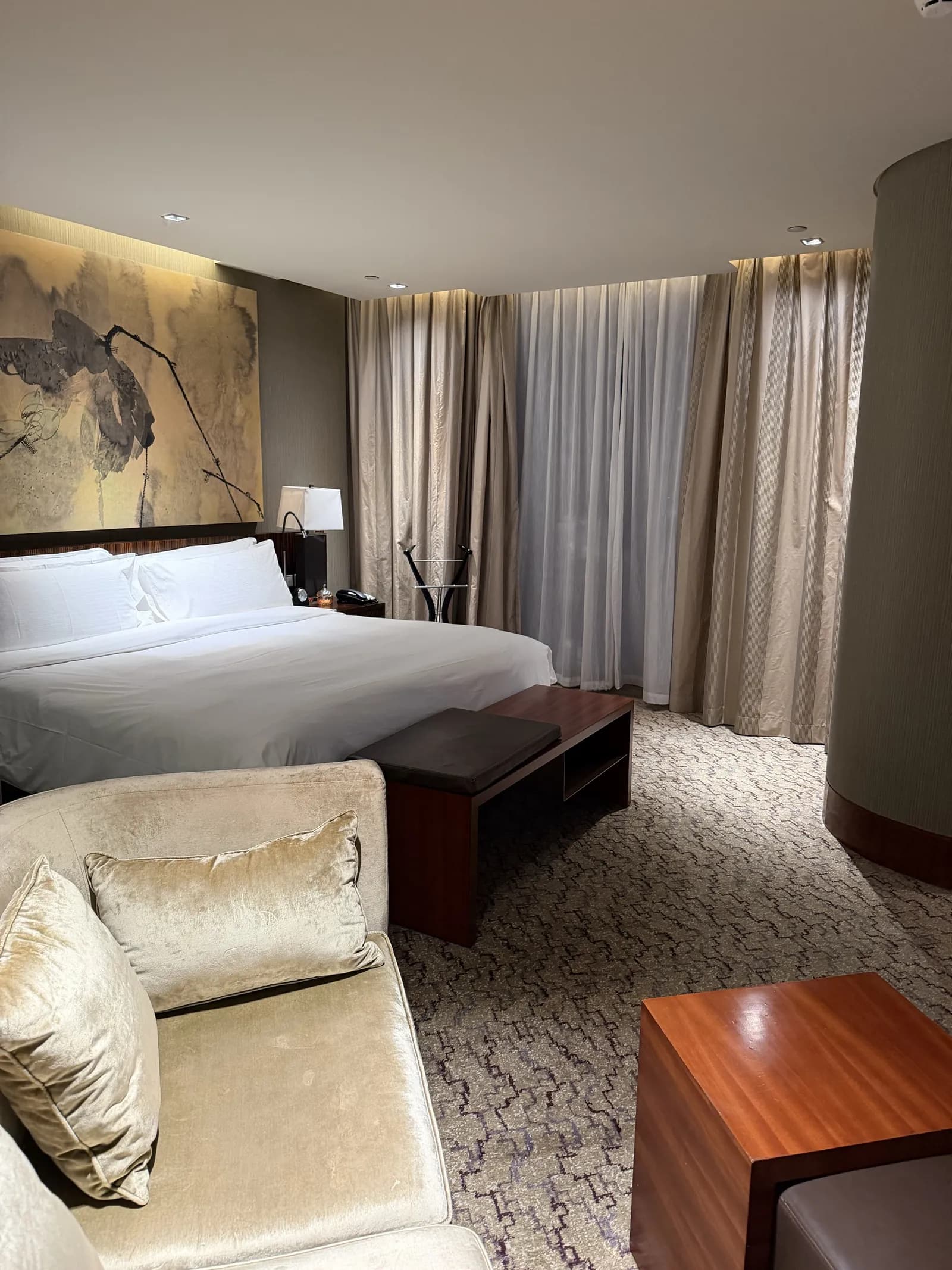 Hotelzimmer mit großem Doppelbett, beigem Samtsofa, hellen Vorhängen und einem Wandbild mit Tuschemalerei