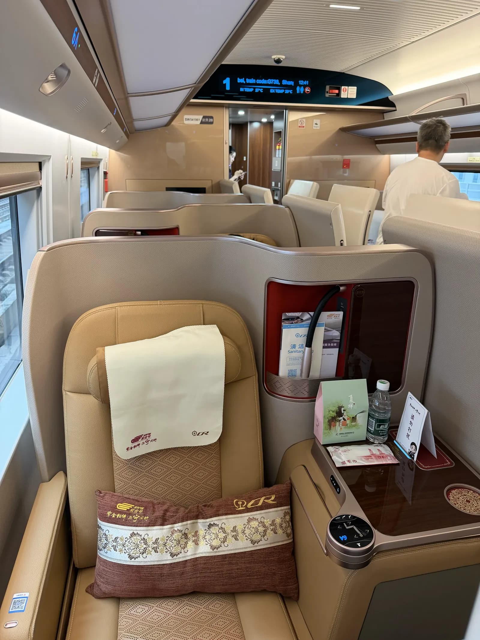Beiger Business-Class-Sitz mit Stoffbezug an der Kopfstütze und einem gemusterten Kissen, daneben eine Ablage mit Wasserflasche, kleiner Box und einem Aufsteller