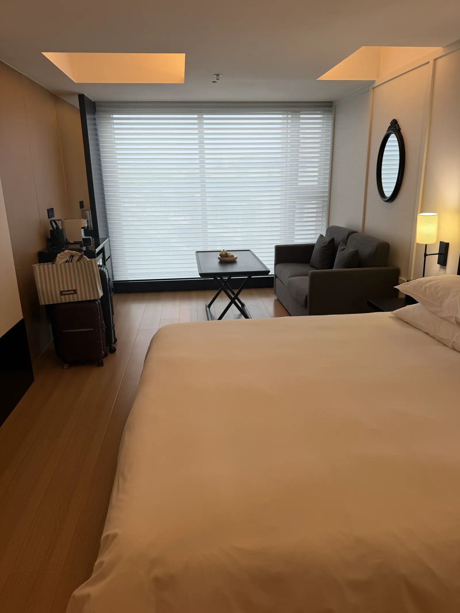 Hotelzimmer mit großem Doppelbett, grauem Sofa vor einem breiten Fenster mit Jalousie, rundem Spiegel an der Wand und Holzboden