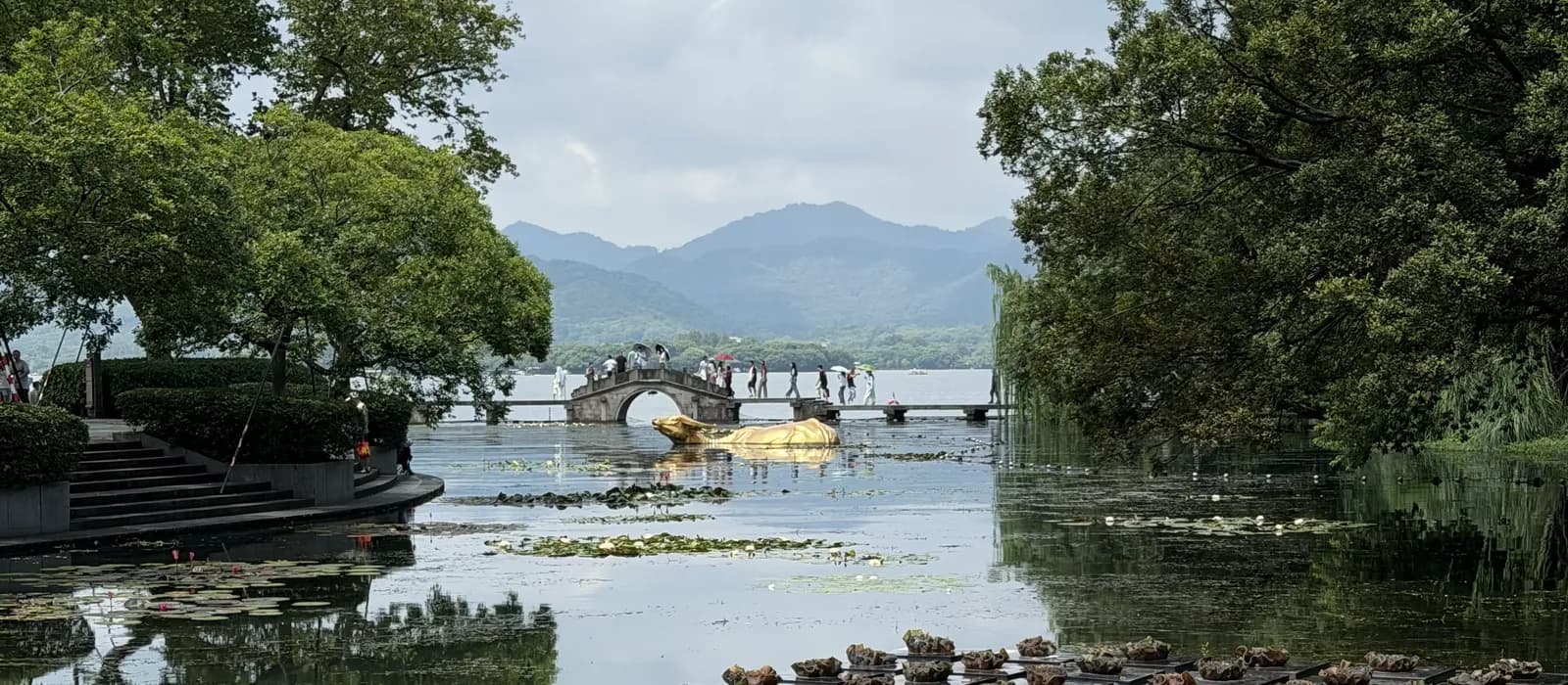 Westsee in Hangzhou mit Seerosenblättern im Vordergrund, einer steinernen Bogenbrücke und bewaldeten Bergen im Hintergrund