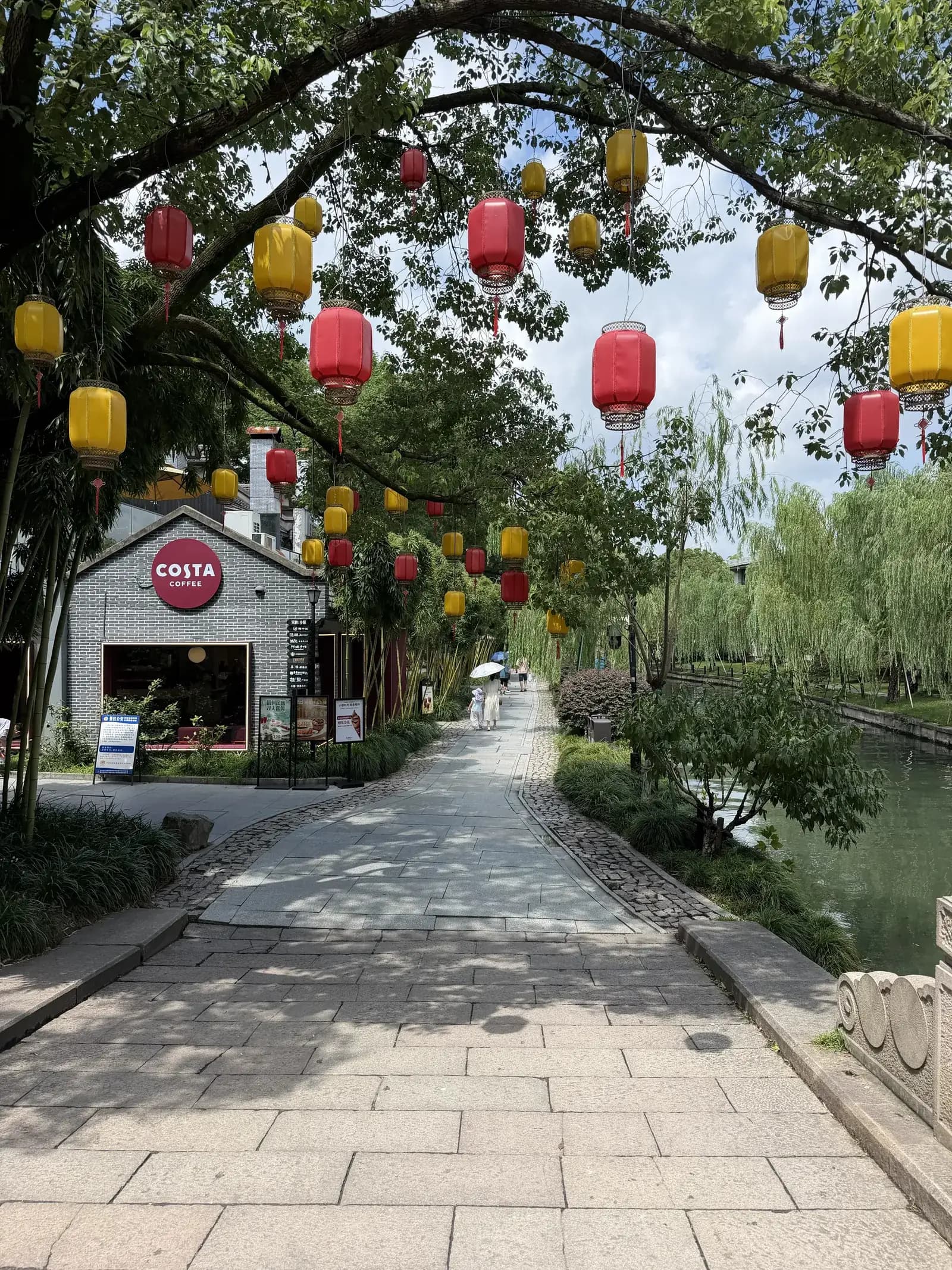 Gepflasterter Weg am Westsee bei Xihu Tiandi mit roten und gelben Laternen in den Bäumen und einem Café in einem Gebäude im traditionellen Stil