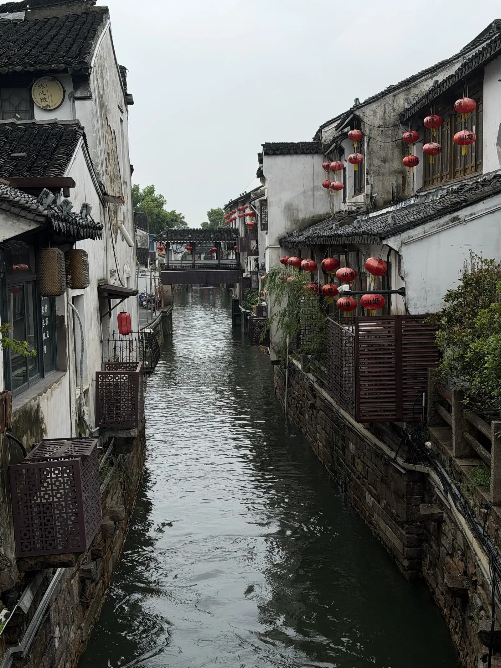 Schmaler Kanal in der Altstadt von Suzhou zwischen weiß getünchten Häusern mit dunklen Ziegeldächern und roten Laternen, im Hintergrund eine überdachte Brücke