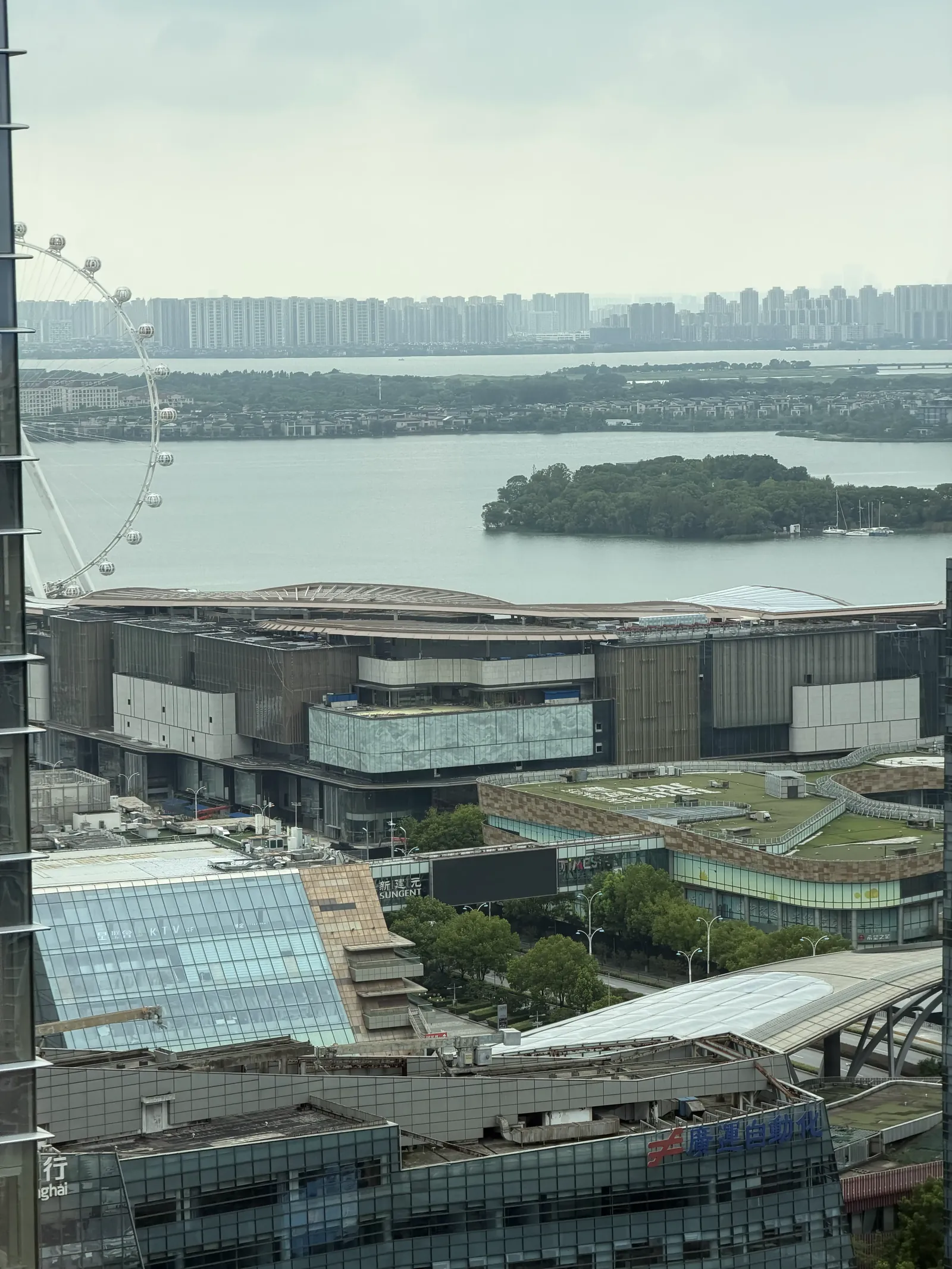 Blick von oben über den Jinji-See in Suzhou mit einer bewaldeten Insel, links ein Riesenrad, vorne ein großer Gebäudekomplex, am Horizont Hochhäuser
