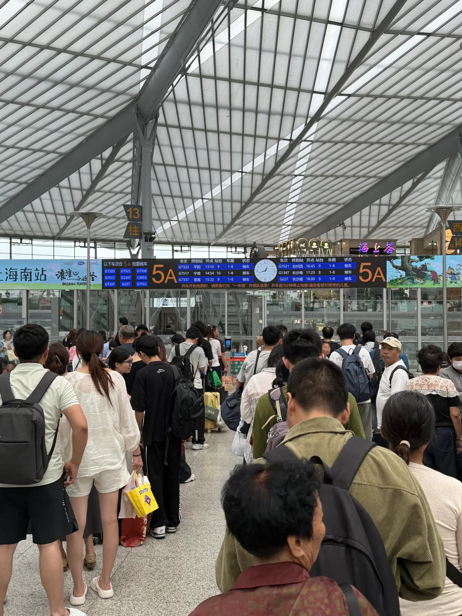 Große Wartehalle im Shanghai South Bahnhof mit Menschen vor dem Gate 5A, zwei Anzeigetafeln mit den nächsten Zugverbindungen, Glasdachkonstruktion, Bereich 上海南站 sichtbar