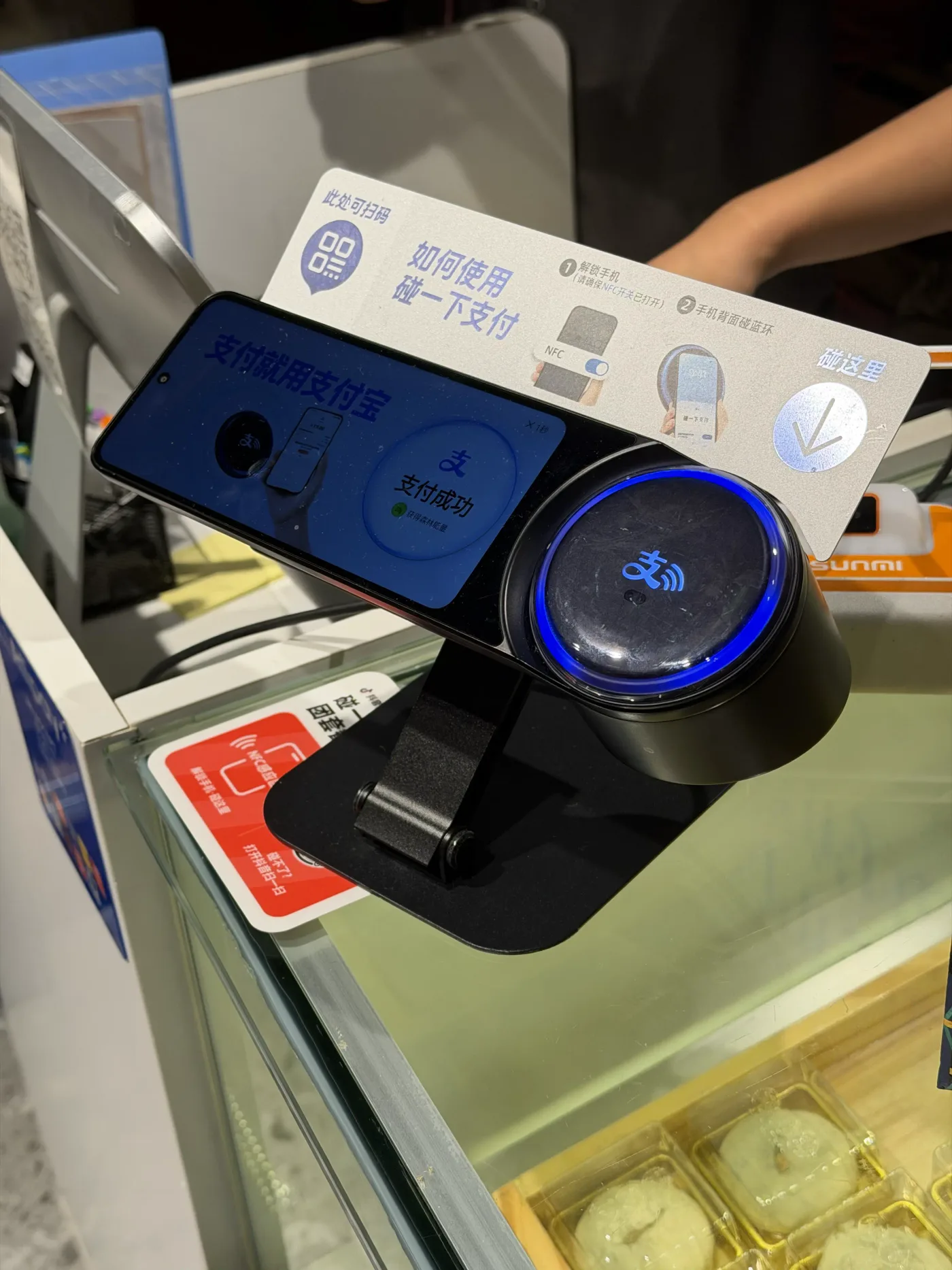 Alipay-Tap-Terminal mit blau leuchtendem Ring an einer Kasse, daneben ein Schild mit der Anleitung zum kontaktlosen Bezahlen